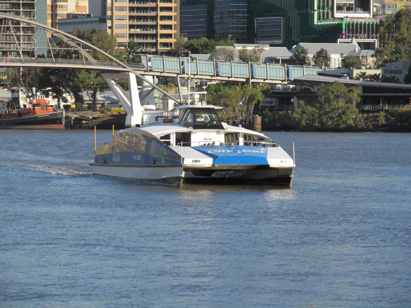 A CityCat ferry on the Brisbane River with buildings and a pedestrian bridge behind it