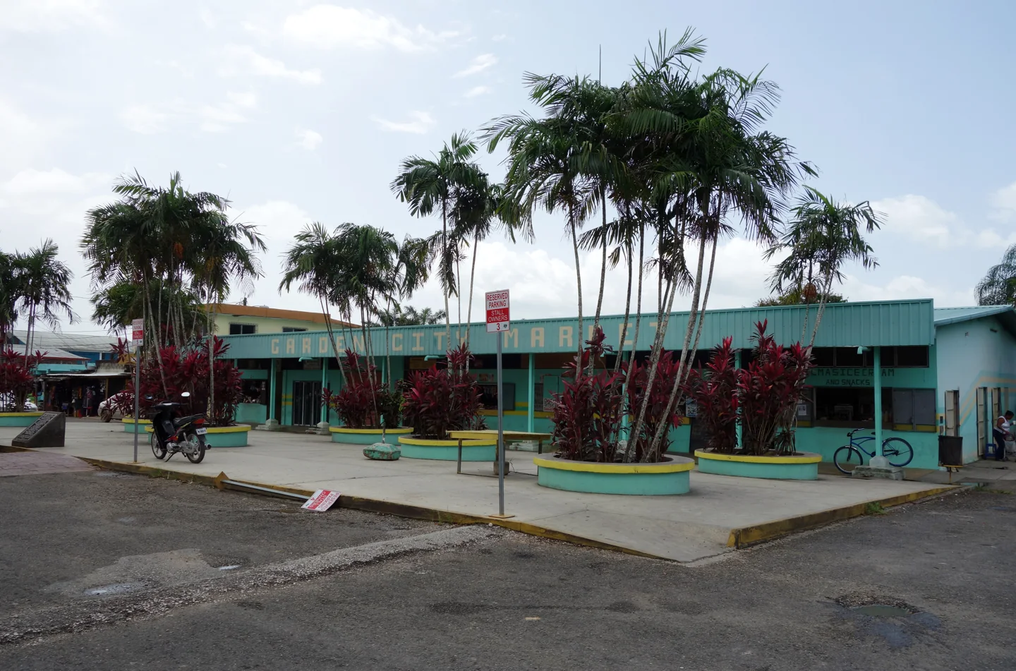 The low turquoise Garden City Market building in Belmopan behind palms and planted forecourt beds.