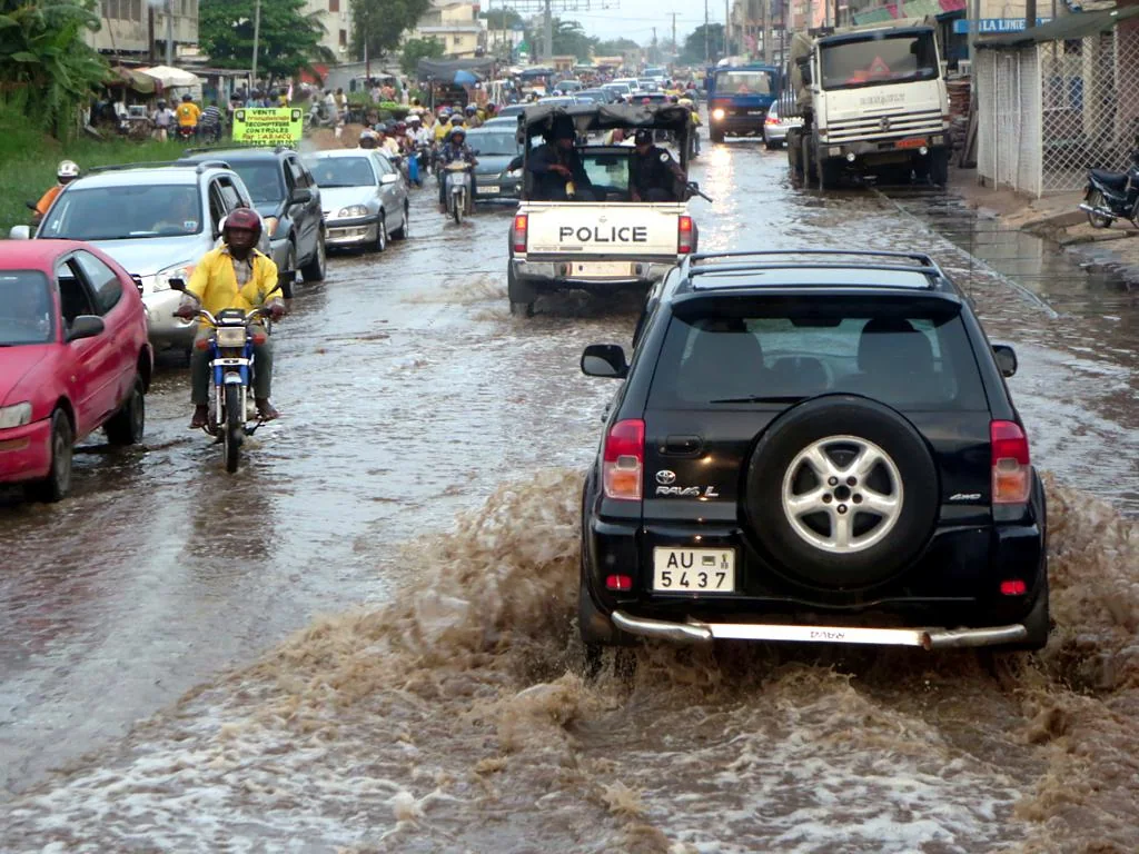 Cars and motorcycles drive through standing water on a Cotonou street.