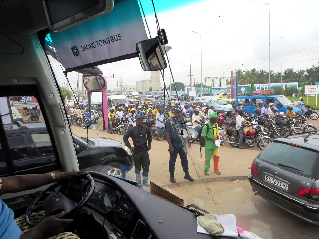 Motorcycles and cars crowd a Cotonou road viewed through a bus windscreen.