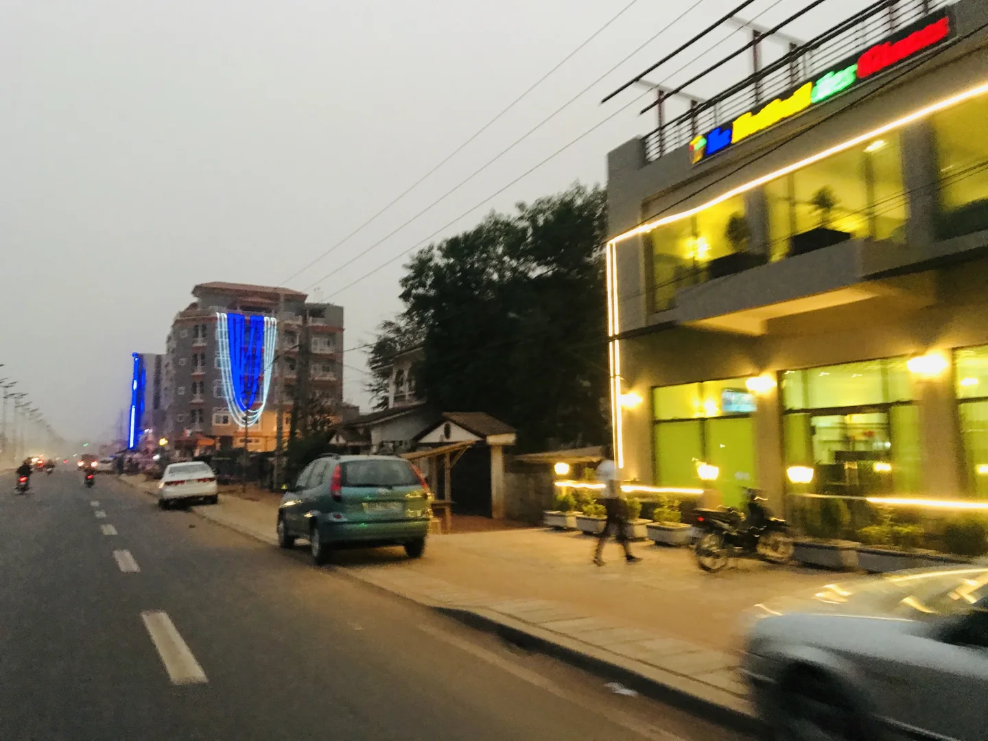 Cars and motorcycles pass illuminated shops on a Porto-Novo street at dusk.