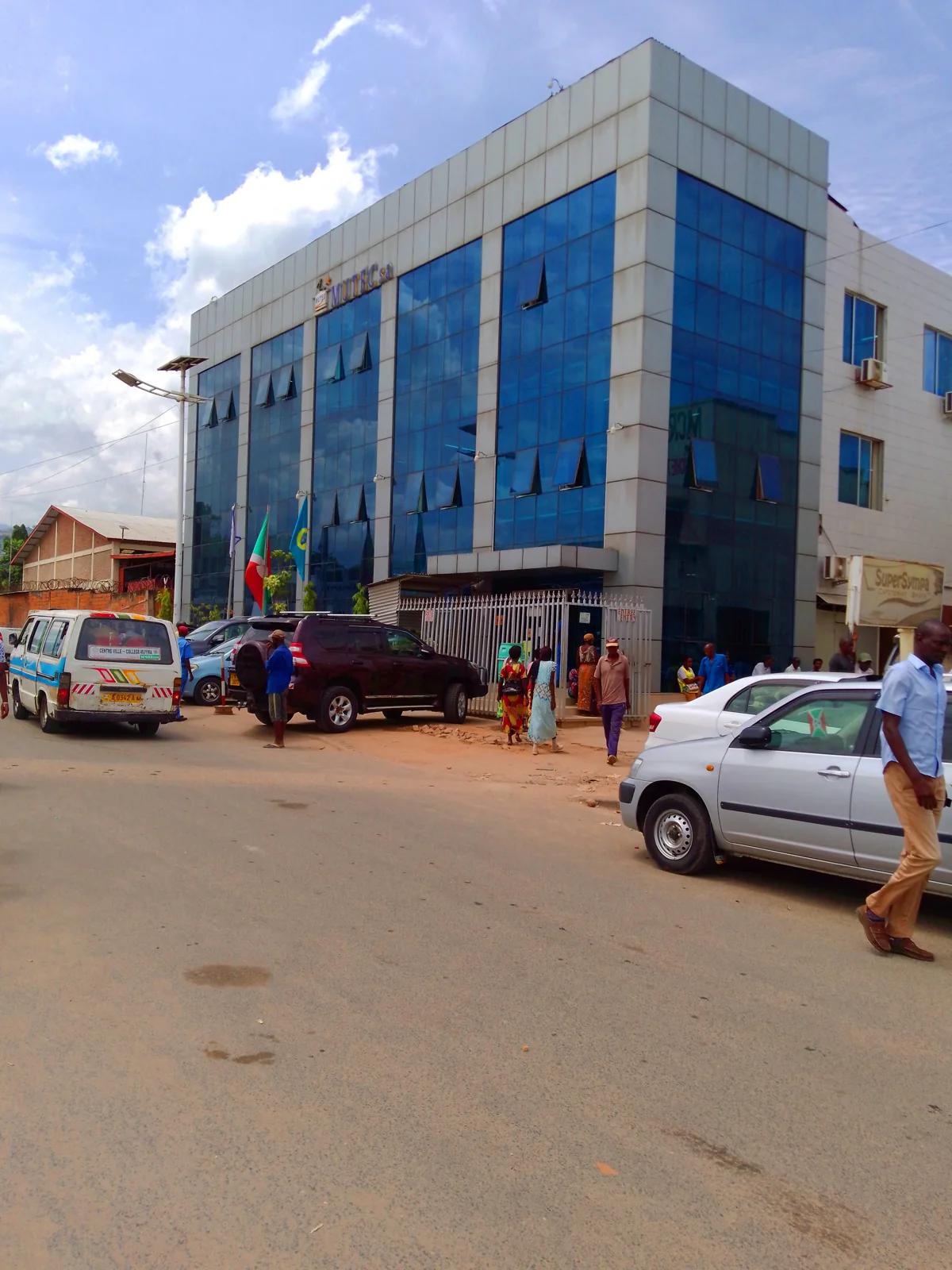 People, a minibus and cars outside a blue-glass commercial building in central Bujumbura