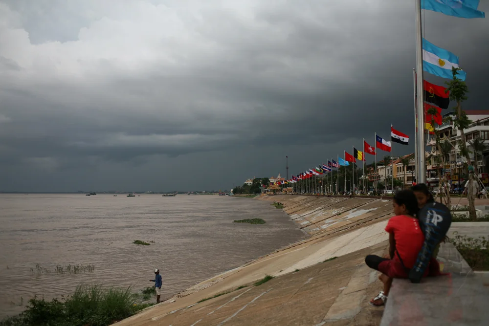 People seated on Phnom Penh’s riverside embankment below a row of flags