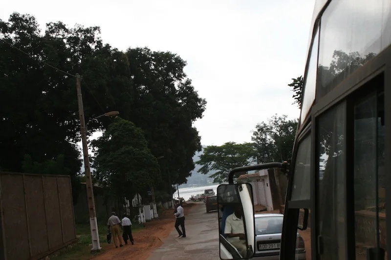 Tree-lined Bangui street seen beside a vehicle, with pedestrians and the river ahead